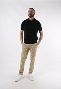 Polo shirt nero a maniche corte, chinos beige e sneakers bianche. Il modello è in piedi con le mani in tasca contro uno sfondo semplice.
