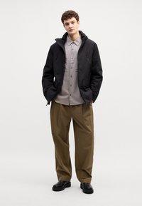 Giacca impermeabile nera con zip, camicia grigia chiara a bottoni, pantaloni beige oversize e stivali neri. Materiali lisci e vestibilità comoda.