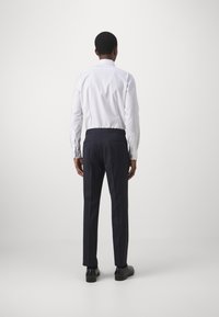 Weißes Langarmhemd mit klassischem Kragen, kombiniert mit dunkelblauen Hosen, die eine taillierte Passform und zwei Gesäßtaschen aufweisen. Sichtbare schwarze Schuhe.