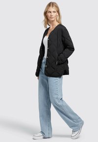 Giacca trapuntata nera con bottoni a pressione, maniche lunghe e vestibilità comoda, abbinata a jeans a gamba dritta blu chiaro e sneakers bianche.
