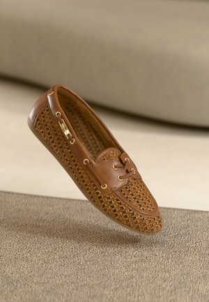 Brun vævet læder loafersko med gyldne snøreringe og en dekorativ guldmetallaccent, der hviler på en tekstureret beige overflade.