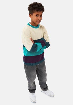 Strickpullover mit cremefarbenen, türkisfarbenen und lila Streifen; hat einen runden Hals und gerippte Bündchen. Kombiniert mit grauen Jeans und weißen Sneakers.