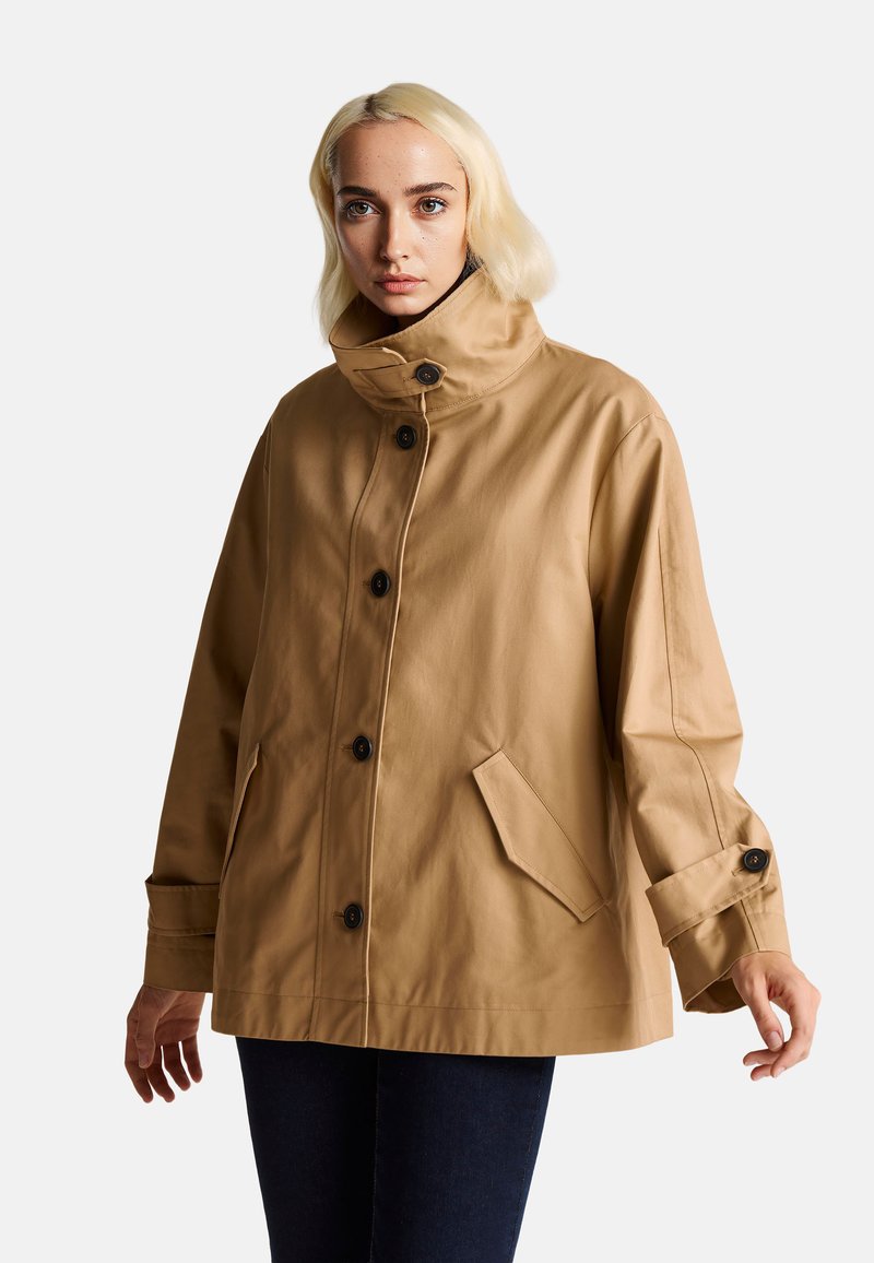 Chaqueta impermeable beige con cuello alto, botones negros, bolsillos laterales y mangas largas. Textura suave con un corte relajado.