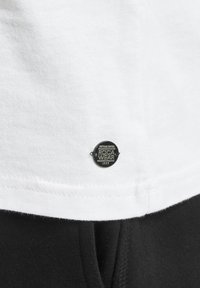 Tricou alb din bumbac cu un eticheta metalică Roca Wear de culoare neagră, având o formă rotundă și o textură netedă, asortat cu un material negru dedesubt.