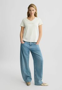 T-shirt crème à col en V avec logo brodé, associée à un jean en denim bleu clair ample et des chaussures dorées à enfiler.