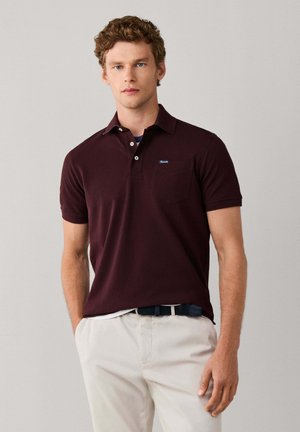 Joven con cabello rizado que lleva una camiseta polo de manga corta de color burdeos oscuro y pantalones beige claros, de pie frente a un fondo liso.