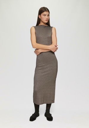 Lang, getailleerd, mouwloos jurkje van een glanzende, brown stof met een gladde textuur. Zwarte panty en platte schoenen complementeren de outfit.