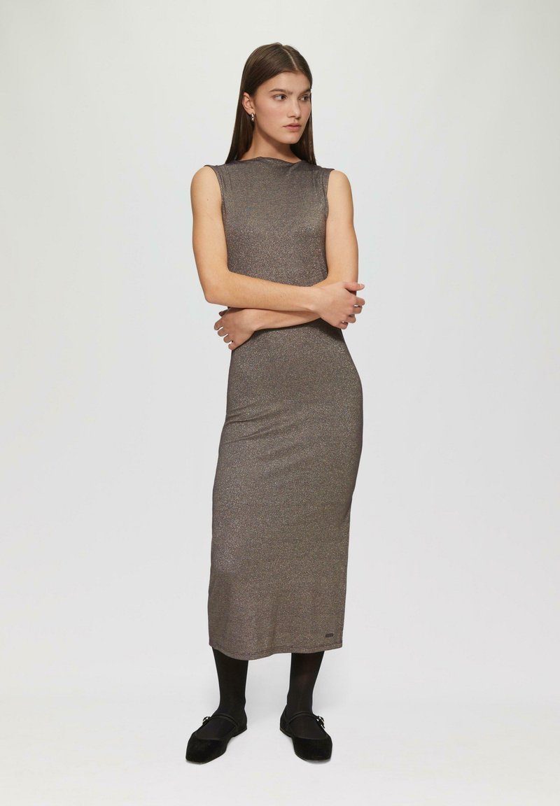 Długa, dopasowana, bezrękawna sukienka z błyszczącego brązowego materiału o gładkiej strukturze. Czarne rajstopy i płaskie buty dopełniają stylizację.