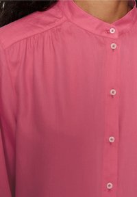 Blusa rosa con colletto alto, chiusura frontale a bottoni e piccoli bottoni. Il tessuto ha una consistenza liscia e morbida con leggere pieghe sulle spalle.