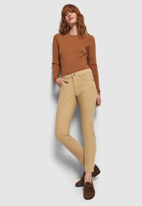 Mujer vestida con un suéter marrón de canalé, pantalones skinny beige y mocasines marrones con estampado de leopardo, de pie con una mano en el bolsillo frente a un fondo blanco.