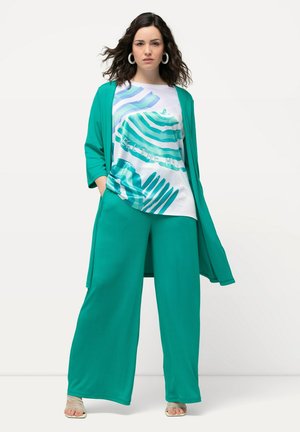 Cardigan lungo leggero color teal indossato su una maglietta bianca a righe blu e con testo. Pantaloni palazzo abbinati e sandali con cinturino.