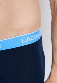 Boxer-briefs navy avec une ceinture élastique bleu clair portant le logo "LACOSTE" en blanc. Texture lisse, design ajusté et sans motifs visibles.