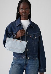 Τζάκετ από τζιν με πορτοκαλί ραφές, λευκή μπλούζα, κρατώντας μια ριγέ μπλε και λευκή τσάντα με καφέ λεπτομέρειες και σκούρους χεριές.