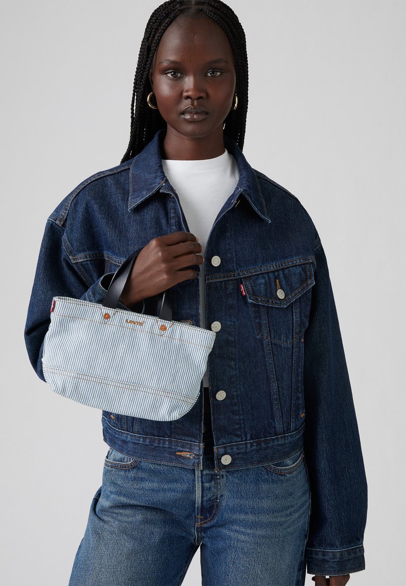 Jeansjacke mit orangefarbenen Nähten, weißes T-Shirt, hält eine blau-weiße gestreifte Tasche mit braunen Akzenten und dunklen Griffen.