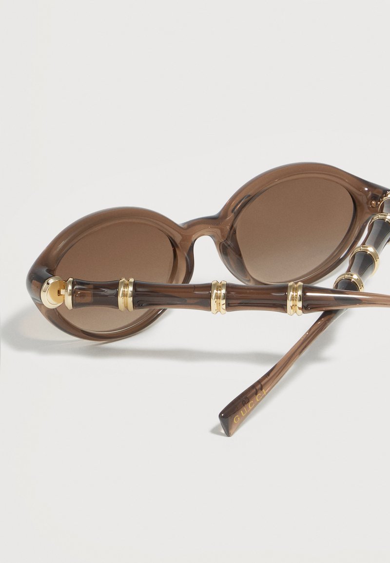 Occhiali da sole rotondi marroni con lenti sfumate e aste in stile bambù accentuate da anelli dorati, posati su una superficie bianca.