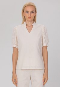 Haut beige clair avec un motif en œillets, manches bouffantes courtes et décolleté en V. Présente un col légèrement volé pour une texture supplémentaire.