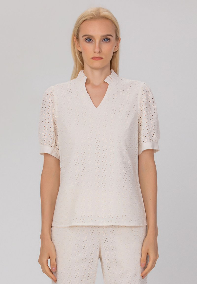 Haut beige clair avec un motif en œillets, manches bouffantes courtes et décolleté en V. Présente un col légèrement volé pour une texture supplémentaire.