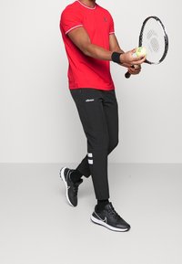 Mies punaisessa paidassa, mustissa verkkareissa ja mustissa tennareissa, pitää tennismailaa ja palloa, valmistelemassa syöttöä yksivärisellä taustalla.