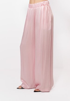 Voľné, široké nohavice s vysokým pásom z ružového saténu s elastickým pásom, nosené s čiernymi sandálmi s otvorenou špičkou, ktoré odhaľujú prsty na nohách.