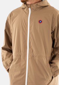 Veste légère beige à capuche avec des poignets élastiques, fermeture éclair blanche et un logo circulaire rouge et bleu sur la poitrine. Texture lisse.