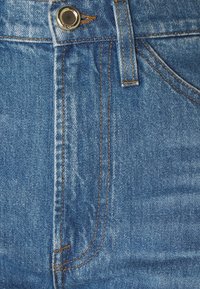 Tejido de mezclilla en azul medio con costuras naranjas en contraste. Presenta un botón de metal y un diseño de bolsillo frontal, superficie texturada con ligero desgaste.