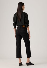 Jeans neri con vita alta e gambe dritte, abbinati a una camicia nera con bottoni. Cuciture notevoli sulle tasche posteriori e una patch in pelle.