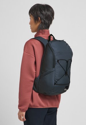 Zwarte rugzak met een gestructureerd oppervlak, verstelbare schouderbanden en bungee koord details. Draag over een rood gewatteerd sweater, zijaanzicht.