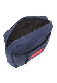 Bolso de tela azul marino con cierre de cremallera, que cuenta con un bolsillo interior y una etiqueta roja en el lateral. Material texturizado y diseño compacto.