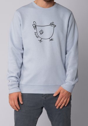 Hombre con sudadera azul clara que lleva un dibujo sencillo en línea negra de un pollo y el texto "le poulet" en el pecho.