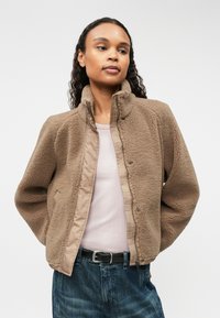 Chaqueta de forro polar en marrón claro con cremallera frontal, cuello alto y bolsillos laterales, llevada sobre una blusa rosa y combinada con jeans azules.