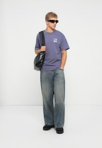 Joven con gafas de sol, camiseta gráfica morada, jeans de pierna ancha, zapatos negros, sosteniendo una gran bolsa negra de hombro, de pie contra un fondo blanco.