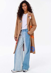 Žena s dlouhými kadeřavými vlasy má na sobě béžový kabát s barevnými nášivkami, bílý tričko, dvoubarevné široké džíny a bílé tenisky, stojí s rukou v kapse.