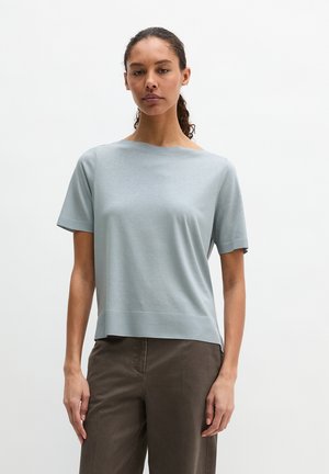 Frau mit zurückgebundenen lockigen Haaren, die ein hellblaues kurzärmliges Oberteil und dunkelbraune Hose trägt, steht vor einem schlichten weißen Hintergrund.