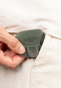 Zelená kožená peněženka s překlopným designem, zdobená bílým prošíváním a kovovým patentním uzávěrem, která je vkládána do světlé kapsy.