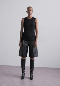 Svart ermeløs topp med vertikale linjer, kombinert med svarte skinnshorts og spisse støvler til knærne. Glatte teksturer, tettsittende design.