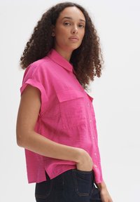 Camicia a maniche corte con colletto di un vivace rosa, in tessuto strutturato, con due tasche anteriori e una vestibilità comoda, abbinata a jeans scuri.