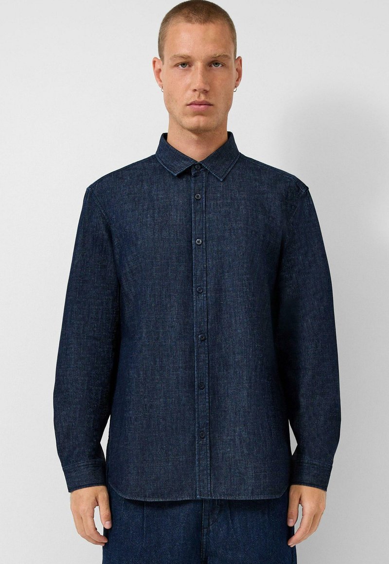 Muž s krátkými vlasy, oblečený v temně modré džínové košili zapnuté na knoflíky a odpovídajících džínových kalhotách, stojící před jednoduchým světlým pozadím.