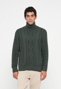 Homem com um suéter de gola alta em malha canelada verde escura e calças bege claras, de pé contra um fundo branco simples.