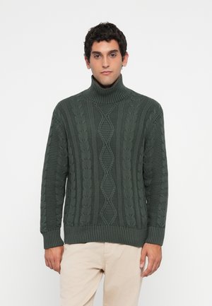 Man wearing a dark green cable knit turtleneck sweater and light beige pants, standing against a plain white background.