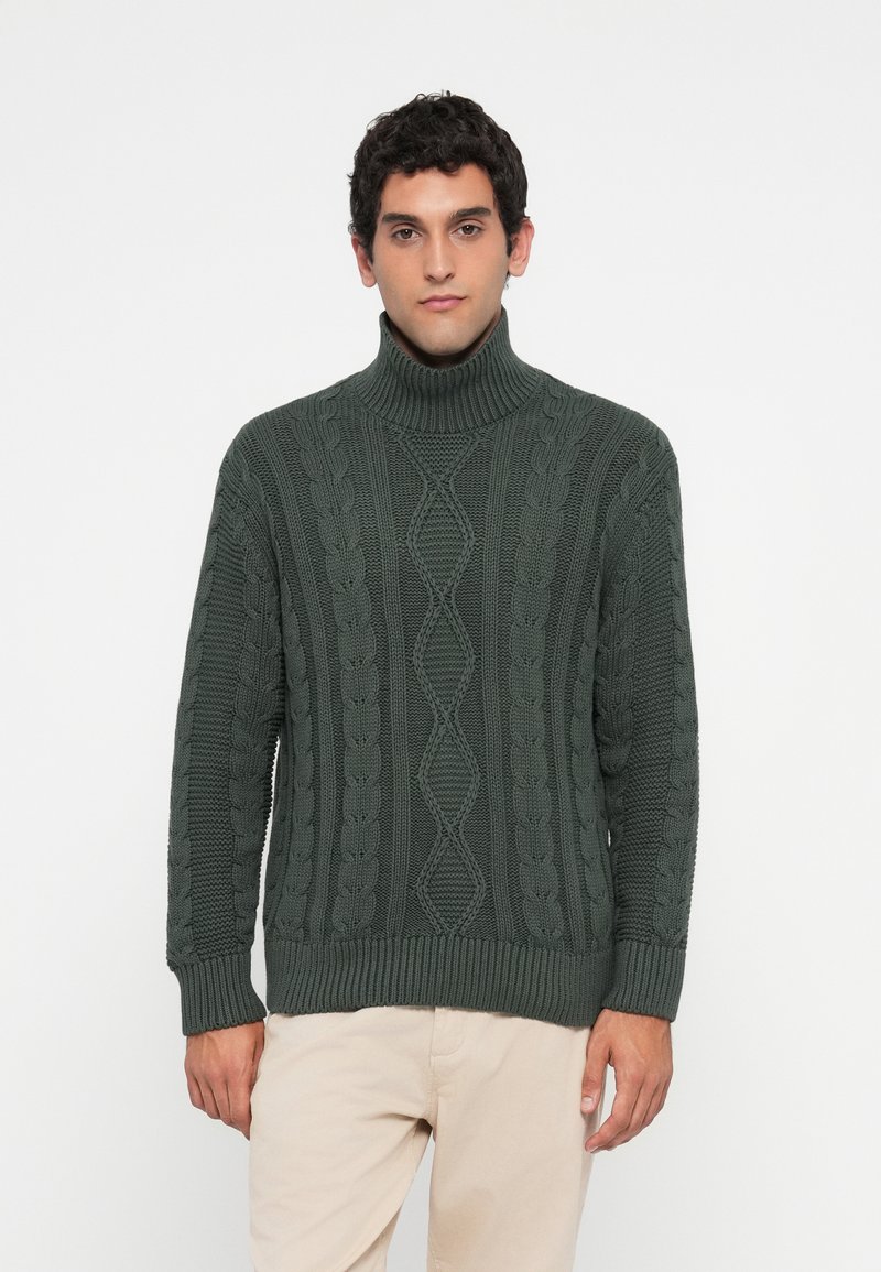 Homem com um suéter de gola alta em malha canelada verde escura e calças bege claras, de pé contra um fundo branco simples.