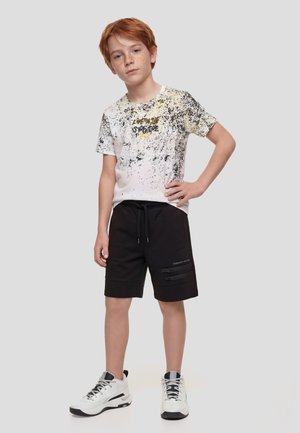 Chico pelirrojo con camiseta blanca con diseño gráfico, pantalones cortos negros con cremalleras y zapatillas blancas, de pie con una mano en la cadera.