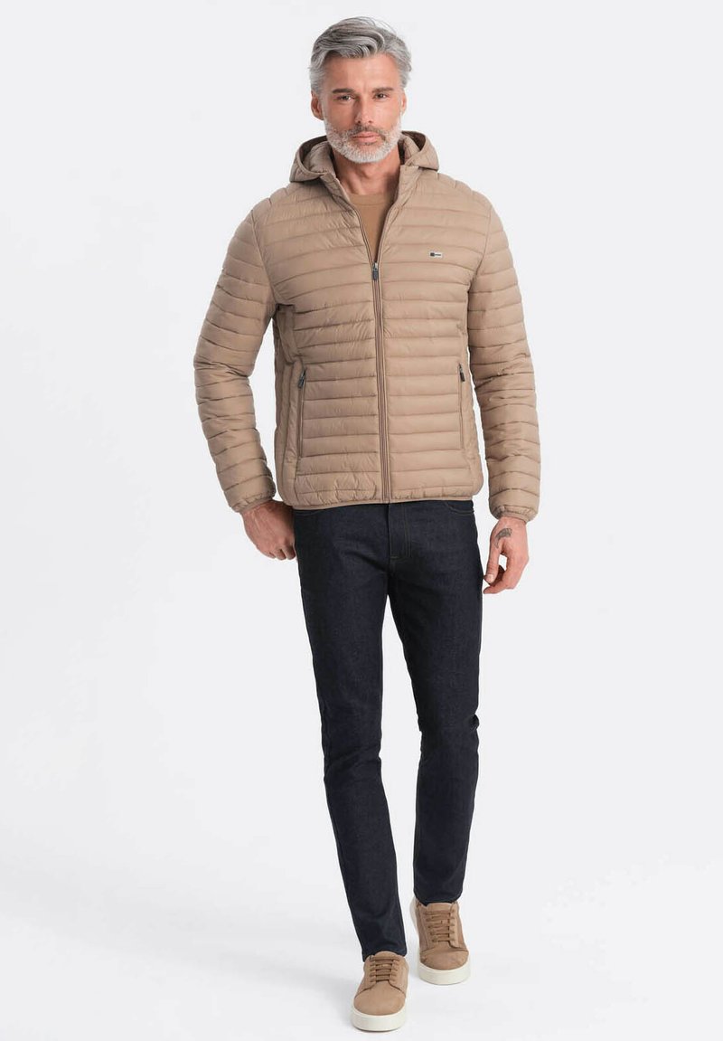 Beige gepolsterte Jacke mit Kapuze, Reißverschluss und Seitentaschen. Über einem braunen Oberteil getragen, kombiniert mit dunklen Jeans und beigen Turnschuhen.