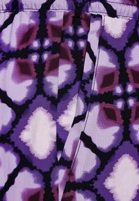 Paarse stof met een geometrisch patroon in tinten van lavendel, donkerpaars en zwart. Gladde textuur met geplooide details.