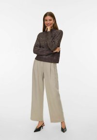 Maglione strutturato di un viola scuro abbinato a pantaloni beige chiaro a gamba larga e mule nere con tacco. La modella si propone in una posa sicura.