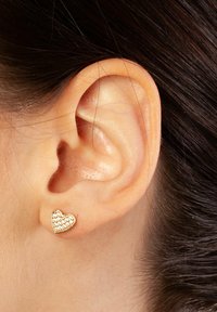 Złote sercowe kolczyki w kształcie serca, pokryte powierzchnią małych, przezroczystych kamieni szlachetnych, prezentowane na małżowinie ucha modelki z ciemnymi włosami.