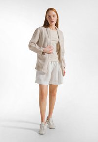 Lekki beżowy lniany żakiet na jasnobeżowej bluzce, zestawiony z białymi lnianymi szortami i lekkimi tenisówkami. Prosty, swobodny fason z podwiniętymi rękawami.