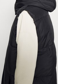 Schwarze Steppweste mit hohem Kragen und Seitenschlitzen, getragen über einem cremefarbenen Langarmkleid. Der Stoff hat eine glatte, glänzende Textur.
