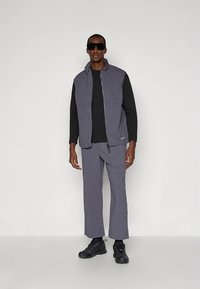 Veste matelassée grise portée sur une chemise noire à manches longues, associée à un pantalon gris assorti. Le mannequin porte des lunettes de soleil noires et des chaussures de sport.