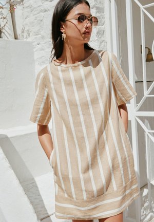 Femme portant une robe beige et blanche à rayures verticales, manches courtes, avec poches, boucles d'oreilles dorées et lunettes de soleil, debout contre un mur et une rambarde blancs.