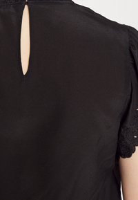 Blouse noire avec une petite ouverture verticale en forme de trou dans le dos et une bordure en dentelle sur les manches courtes, portée sur un bras à la peau claire.